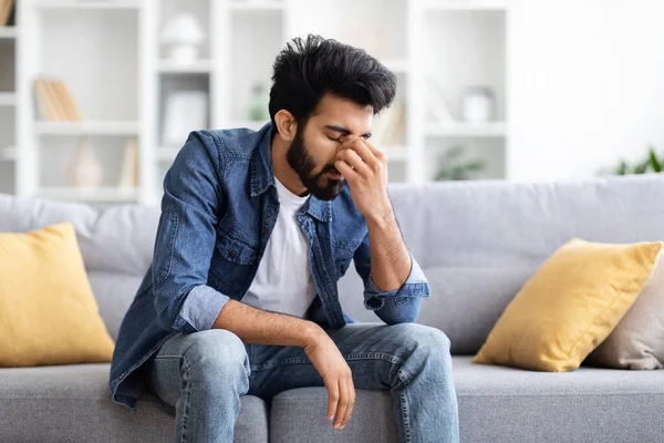 Seasonal Depression. Portrait Of Sad Young Indian Man Sitting On Couch At Home, Upset Tired Eastern Guy Resting In Living Room, Suffering Life Problems Or Mental Breakdown, Copy Space