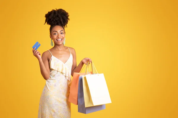 グラッドペンシブな若い黒人女性はクレジットカードを見せ 購入を夢見て 黄色の背景 スタジオで隔離された購入で多くのパッケージを保持します アドバイスとオファー ライフスタイル ショップホリックのための販売 — ストック写真
