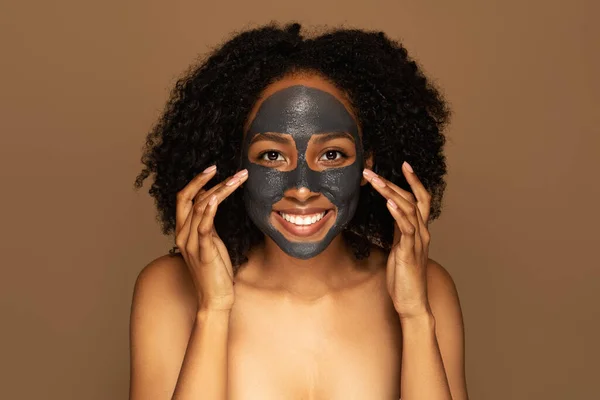 Cheerful attractive half-naked young black woman with purifying anti-blemish black mask on her face isolated on brown studio background. Face care, beauty routine concept