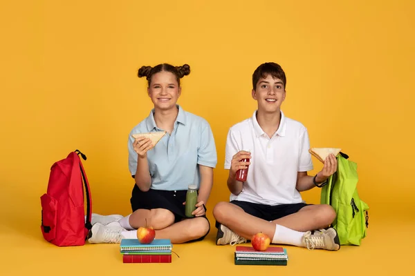 快乐的少年欧洲男孩和女孩背着背包吃苹果和三明治 喝果汁瓶 隔离在黄色的工作室背景 友谊和午餐 学校教育 休息吃饭 — 图库照片