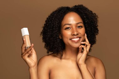 Mutlu, çekici, yarı çıplak, parlak tenli, güzel ürün temeli ya da kapatıcısı olan, kahverengi stüdyo arka planında izole edilmiş bir şişe gösteren siyahi bir kadın.