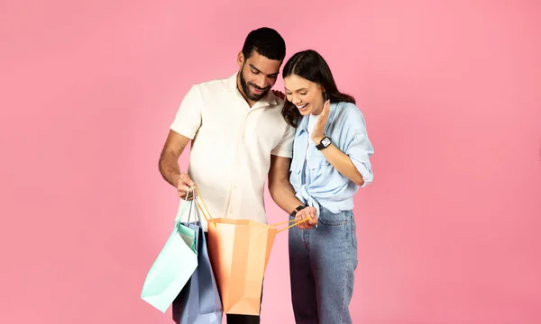 カラフルな紙袋の中を見ている幸せなかなり若い女性 ブラックフライデー中に彼女のボーイフレンドや夫と買い物 ピンクの背景に隔離されたシーズンセール コピースペース 小売り 消費者主義 — ストック写真