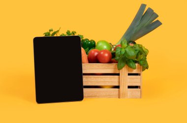 Üzerinde taze sebzeler ve meyveler olan tahta kutunun yanında duran boş ekranlı dijital tablet. Tarım sitesi Reklamları ya da Çevrimiçi Market Alışverişi İçin Model
