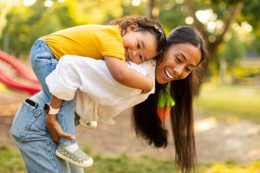 Neşeli Japon annesi, sevimli küçük kızını sırtında taşıyor, oyun sahasında eğleniyor, haftasonu dışarıda Green Park 'ta gülüp poz veriyor. Çocuk ve anne dışarıda eğleniyor.