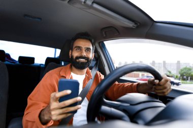 Araç kullanırken seyrüsefer için mobil uygulama kullanan akıllı telefonu tutan mutlu bir Arap şoför. Gps araba navigatör uygulaması kullanarak yeni modern otomobilde gezinti yapmaktan hoşlanan bir adam.