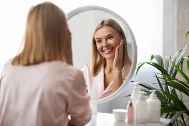 Cilt bakım konsepti. Güzel, orta yaşlı bir kadın, aynaya bakarken yüzüne dokunuyor çekici, olgun bir kadın, güzellik tedavisinden sonra yumuşak teninden zevk alıyor, seçici bir şekilde odaklanıyor.