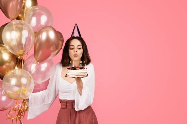 Bin yıllık neşeli beyaz şapkalı esmer kadın pastaya mum üflüyor, pembe arka planda, stüdyoda izole edilmiş. Reklam ve teklif, kutlama tatili, doğum günü partisi