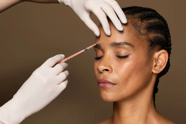 stock image Beautician hands making hyaluronic acid injection for glabella facial rejuvenation procedure, black middle aged woman getting cosmetic injection between eyebrows, brown studio background