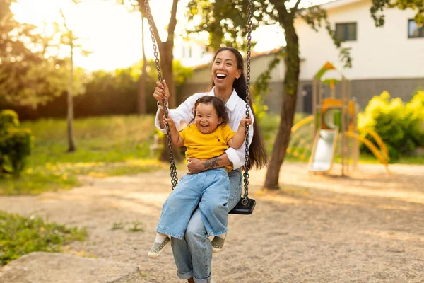 Joyful Asian Mommy And Little Baby Daughter Sitting On Swings Together And Riding Having Fun At Outdoor Playground In Park. Mommy And Infant Enjoying Ride Bonding On Summer Weekend Outside