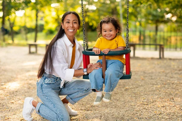 Cheerful Chinese Mom Riding Infant Daughter While Baby Sitting On Swings And Smiling At Camera, Having Fun Together, Posing At Modern Playground Outdoor. Mother And Toddler Spending Weekend In Park