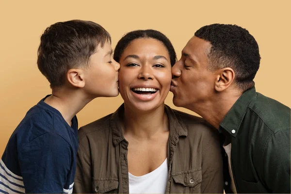 Mothers day. Loving millennial black father and preteen kid son kissing happy woman, showing love and affection, greeting their mother and wife with birthday or womens day, closeup