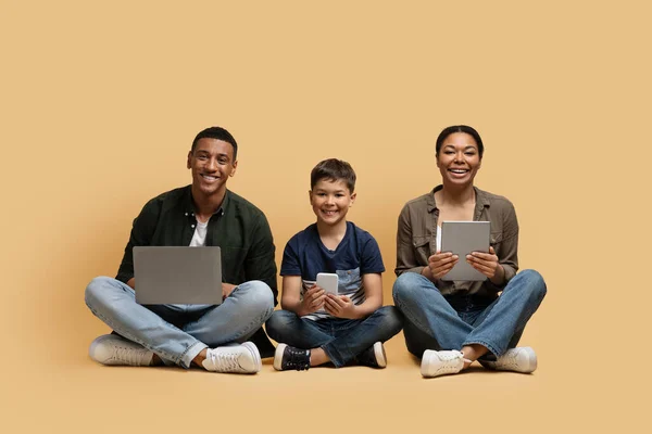 Gadgets addiction. Young black family of three holding and using different electronic devices while sitting on floor on beige background. Parents and their preteen son with modern gadgets