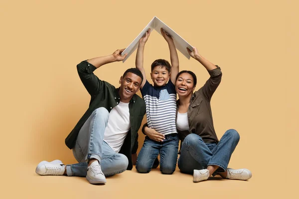 Cheerful loving millennial black parents and their son holding cardboard roof above their heads, father and mother take care about their kid. Cute family moving to new house, beige background