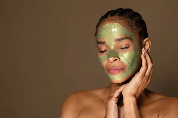 Sensual african american middle aged woman with green peel-off mask on her face posing isolated on brown background, copy space. Modern cosmetics products for aged skin
