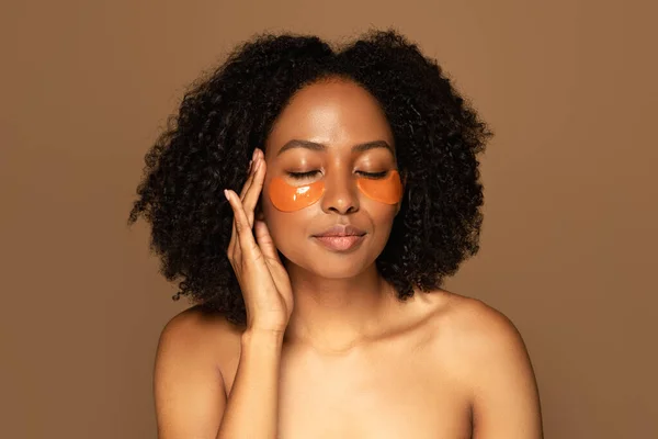 Headshot of attractive topless young black woman with bushy hair and perfect even healthy skin using eye patches, isolated on brown background, copy space. Skin care and spa treatments, web-banner