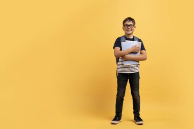 Sarı stüdyo arka planında izole edilmiş, gözlüklü, sırt çantalı ve not defterli neşeli, şirin çocuk okulu öğrencisi. Okul, çocuk eğitim konsepti