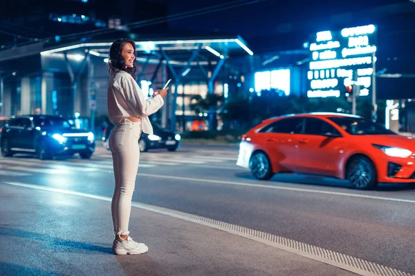Full length shot of young woman standing on the street in the night holding smartphone, waiting taxi, free space. Young lady tracking gps via location app. Busy city traffic