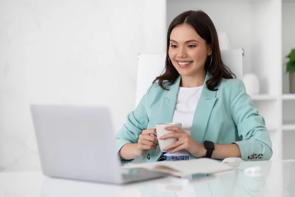令人高兴的是 穿着西服的千年期高加索女士在办公室休息 办公室里装着一杯茶和一台电脑 喝咖啡休息 与设备打交道 经理工作 视频通话和会议 — 图库照片