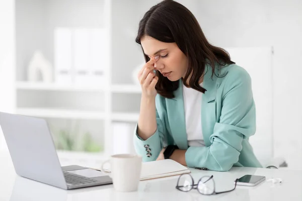 Unhappy tired millennial caucasian lady in suit rest at workplace with computer, suffer from headache in office interior. Problems in business and health, manager work, overwork, deadline and ill