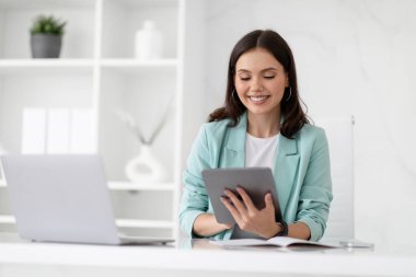 Takım elbiseli neşeli beyaz bayan yönetici iş yerindeki tabletine dizüstü bilgisayarla yazı yazıyor, ofis içi cihazlarla eğleniyor. Çalış, uygula ve müşteriyle uzaktan sohbet et