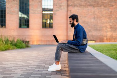 Çevrimiçi teklif. Kentsel alanda dizüstü bilgisayarda yazan Hintli adam, çalışıyor ve web sitesini kullanıyor. Modern Serbest Konsept, İnternet Kariyeri. Guy Websurfing Via Bilgisayar, Side View 'da. Boş alan
