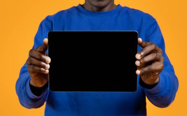 Gündelik kıyafetli siyahi adam yatay modern dijital tablet ve boş siyah ekran gösteriyor. Güzel bir uygulama, online asistan, sohbet robotu, model, sarı stüdyo geçmişi var.