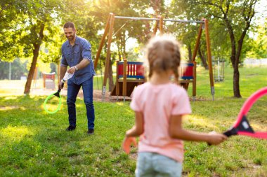 Güneşli bir yaz gününde babasıyla parkta oynarken badminton raketi tutan küçük bir kızın arka görüntüsü.
