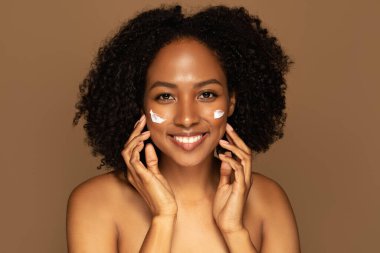 Yüzüne nemlendirici ya da nemlendirici organik güzellik ürünleri kremi sürerek gülümseyen, kahverengi arka planlı, genç, güzel, üstsüz, hoş siyah bir kadın. Cilt bakım rutini