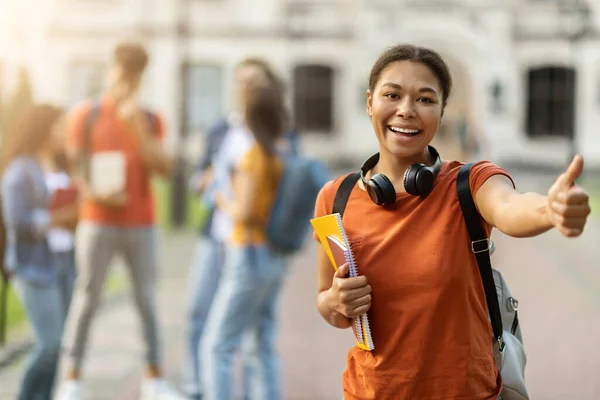 Study Abroad. Happy Young Black Female Student Posing Outdoors And Showing Thumb Up, Cheerful African American Woman Carrying Backpack And Workbooks, Recommending College Program, Selective Focus