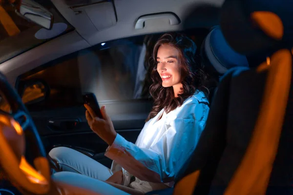 夜は高級車の乗客席に座りながらスマートフォンを使用するハッピーな白人女性 携帯電話を使ってスクリーンを見たり笑ったり — ストック写真