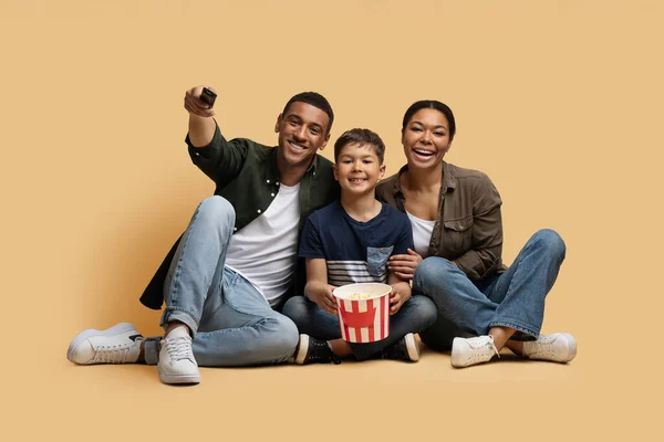 アフリカ系アメリカ人の家族が映画を見たり バケツからポップコーンを食べたり 床にかわいい子供の息子と座っている両親 ベージュの背景に隔離されたリモートコントロールを保持している男 — ストック写真