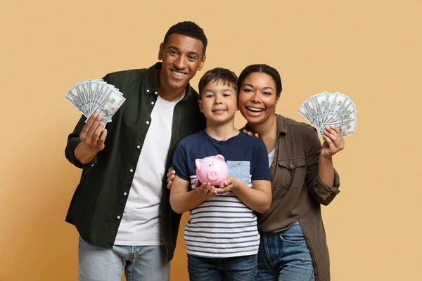 Family savings, budget planning, childrens pocket money. Positive loving young black father mother and preteen kid son holding cash dollar banknotes and piggy bank, isolated on beige background