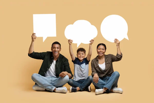 Communication in family. Black mom, dad and son holding different empty speech bubbles above their heads, happy family of three sitting on floor on beige studio background, copy space