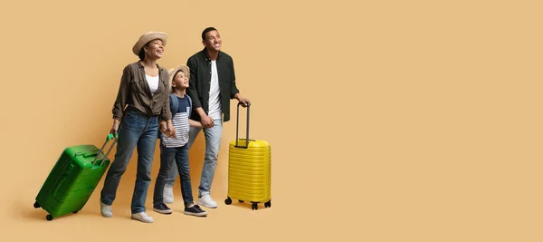 Family vacation, summer trip together concept. Happy young black family tourists travelling together, father, mother and school aged son with suitcases looking at copy space on beige, web-banner