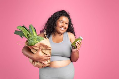 Gülümseyen Afro-Amerikan kadın spor kıyafetleri içinde elinde yeşil sebze ve meyve suyu şişesiyle pembe stüdyo arka planında poz vermiş. Kilo kaybı, vücut ve sağlık hizmetleri