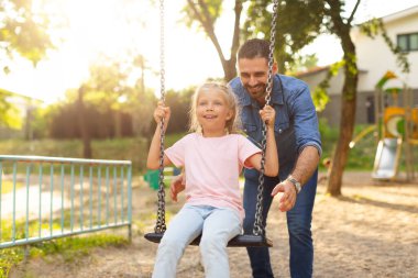 Park maceraları. Mutlu orta yaşlı bir baba, küçük kızıyla oyun bahçesinde salıncaklarla oynuyor, dışarıda eğleniyor. Aile yaşam tarzı konsepti