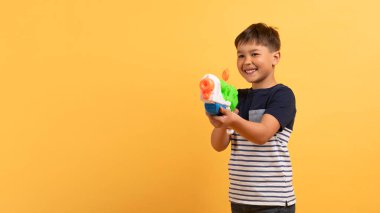 Yaz kıyafetleri içinde küçük mutlu bir çocuk sarı duvar arkasında izole edilmiş oyuncak su tabancası tutuyor. Çocuk stüdyo resmi. İnsanlar çocukluk konsepti, kopyalama alanı olan panorama