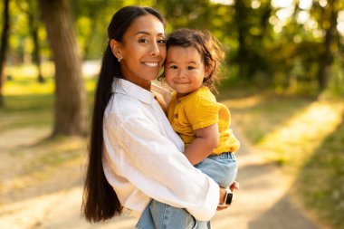 Asyalı anne şefkatle sarılıyor ve sevimli kızını kucaklıyor dışarıda yeşil parkta duruyor kameraya gülümsüyor. Küçük şirin bir çocuğu taşıyan mutlu bir annenin portresi, birlikte gezintiden zevk alıyor.