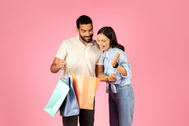 Heyecanlı mutlu genç adam ve kadın pembe arka planda günlük kıyafet alışverişi yapıyorlar. Neşeli alışveriş meraklıları renkli alışveriş torbalarını kontrol ediyor. Perakende kavramı