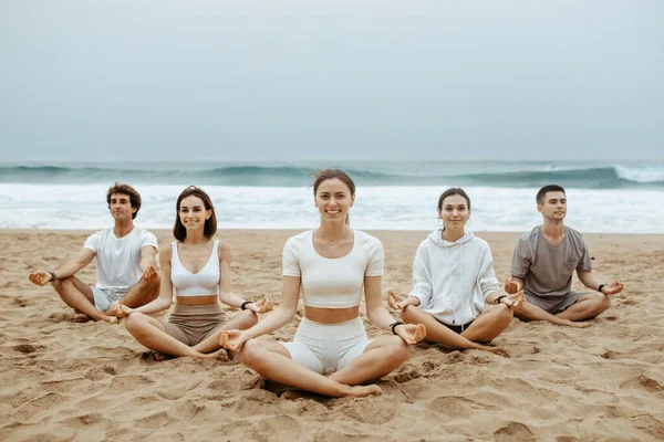 年轻人在海滩瑜伽中找到了平衡点 在沙滩上静坐着荷花 在海滨享受晨练 并对着镜头微笑 — 图库照片