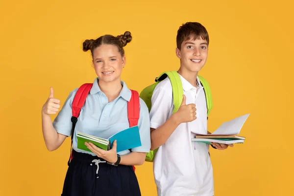 令人高兴的是 聪明的高加索女孩和男孩带着书本 背包露出大拇指 与橙色工作室背景隔离 学习建议 批准学校教育 — 图库照片
