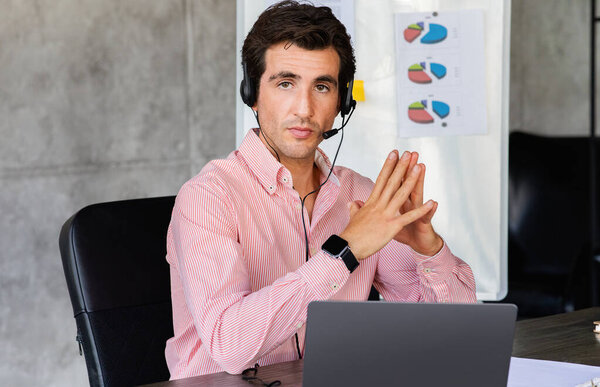 Focused young businessman wearing headphones, holding video call with clients on laptop. Concentrated millennial man giving online educational class lecture, consulting customer, office interior