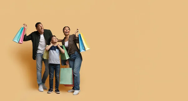 Happy excited black family millennial parents and school aged boy kid enjoying shopping together, holding colorful purchases and smiling, looking at copy space, isolated on beige background