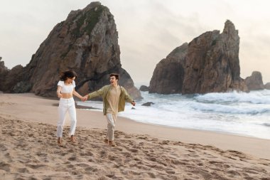 Heyecanlı genç Avrupalı çift el ele tutuşuyor ve okyanus kıyısı boyunca sahilde koşuyor, birlikte eğleniyor ve zaman geçiriyorlar, tam boy, kopyalama alanı.