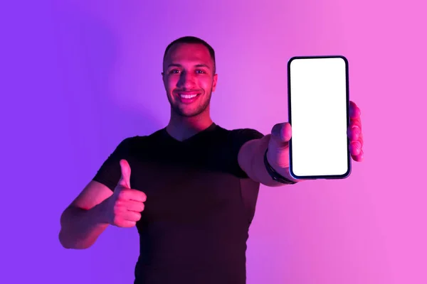 ハッピーブラックスポーツマンは 空の画面と親指で携帯電話を表示し カメラに微笑み トレーニングと減量アプリケーションを推奨し 紫のネオンバックグラウンドにポーズ モックアップ — ストック写真