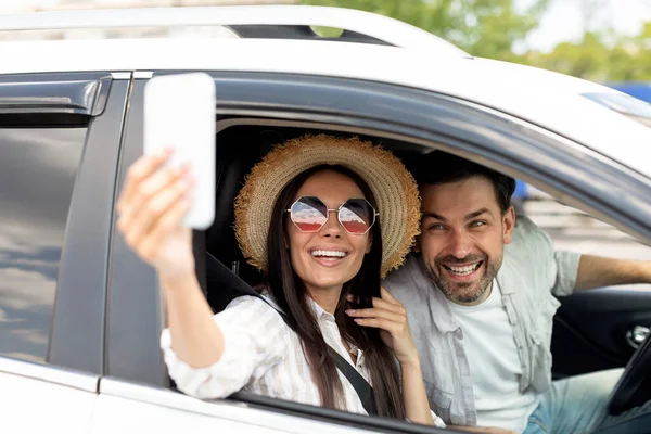 ハッピーな若いカップル30代は 携帯電話を使用して 夏の帽子とサングラスを身に着けている車の中でセルフィーを取ります エキサイティングな陽気なミレニアル世代の男女が休暇に行く 自動車で写真を撮る — ストック写真