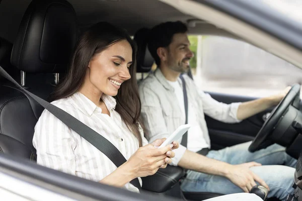 Online reservation. Happy millennial couple sitting inside white car, woman holding smartphone, cheerful man driver looking at road, lady booking hotel online, using travelling application