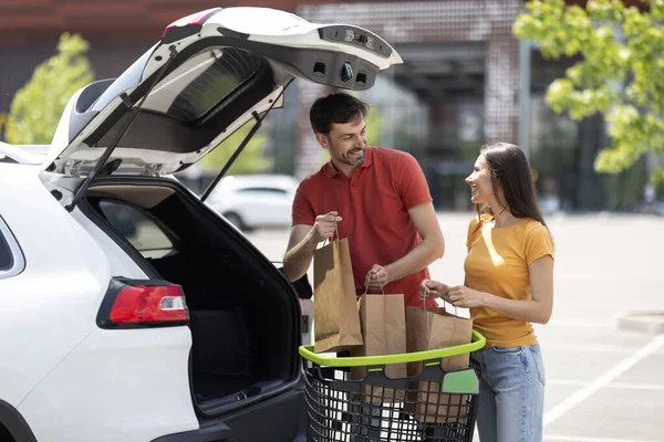 Millennial spouses packing car trunk with groceris. Cheerful loving young man and woman in casual standing by auto with shopping cart full of purchases, going home from supermarket