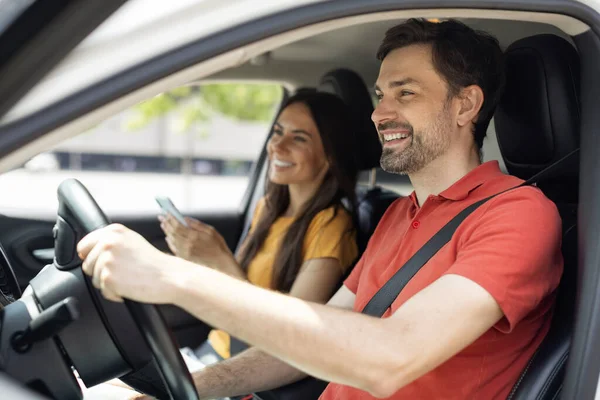 ショート ロード ルート 高級車の中に座っている笑顔のミレニアルカップル スマートフォンを持っている女性 道路を見ている陽気な男性ドライバー デジタルマップアプリケーションを使用して場所を選択する女性の肖像画 — ストック写真