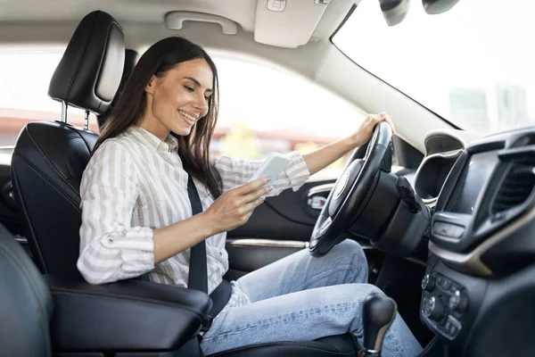 ドライビングコンセプト スマートフォンを運転しながらカジュアルな衣装ドライバーでポジティブな笑顔の若い女性 インターネットでスクロールしたり 自動車に座っている間に友人にテキストを送る女性 — ストック写真